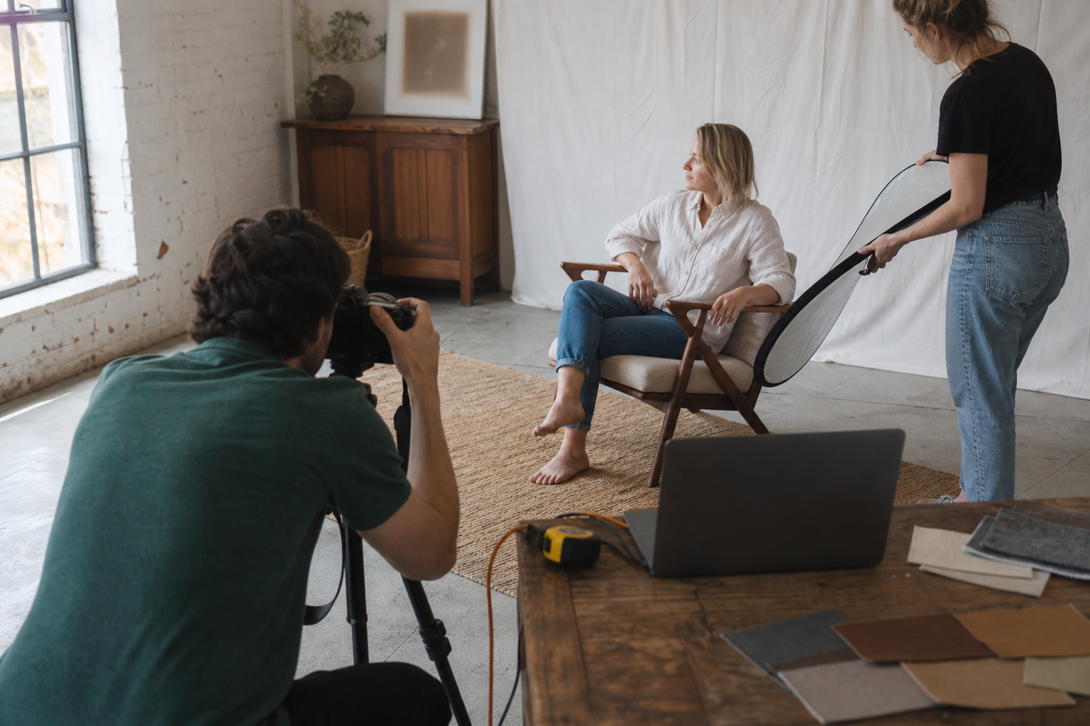 Furniture content team photographing a person seated naturally in a lounge chair in a daylight studio.